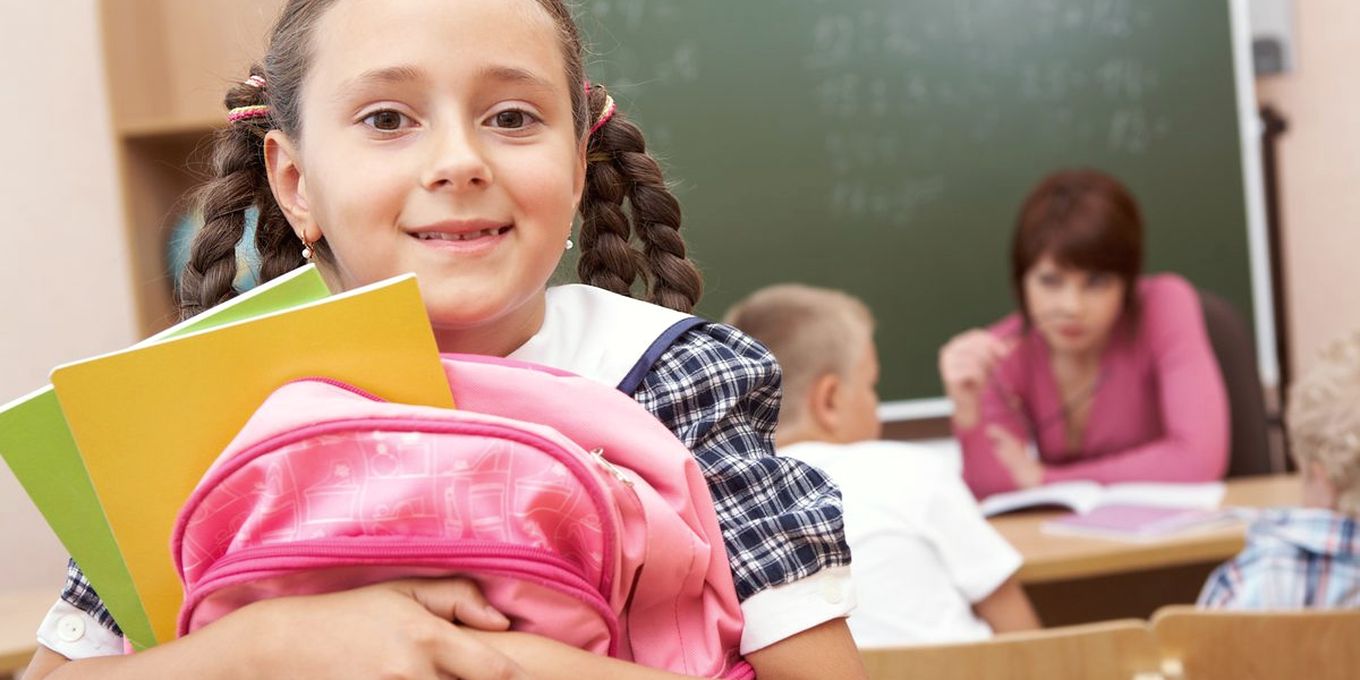 Una alumna abraza su mochila en el aula, con sus compañeros al fondo
